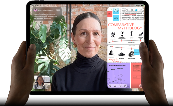 iPad Pro, front exterior, landscape orientation, space black color, rounded corners, black display bezel, hands holding left and right sides, display showing a FaceTime conversation being live translated, and an educational app showing a presentation of "Comparative Mythology" with text, diagrams, and handwritten notes